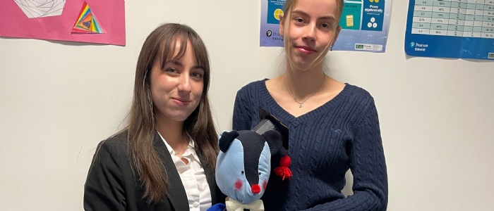 Two female sixth form students smiling and holding their team mascot, a blue teddy bear made of OSH uniform